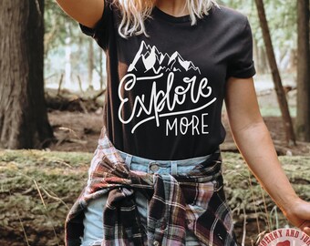 以下が含まれることがあります： 白い山と「Explore More」の文字が書かれた黒いTシャツ。森の中で人が着ています。