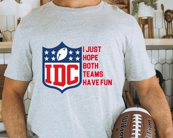 Può includere: Una maglietta grigia con una grafica blu e rossa di un casco da football con stelle e il testo "IDC I JUST HOPE BOTH TEAMS HAVE FUN".