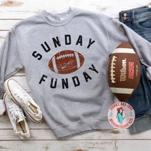 Pu&ograve; includere: Felpa grigia con il testo "SUNDAY FUNDAY" in nero e una grafica di football americano marrone usurata. Un pallone da football americano marrone con lacci bianchi e il testo "Wilson. NCAA OFFICIAL SIZE" si trova nell'angolo in alto a destra dell'immagine.
