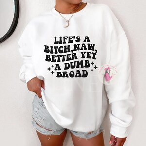 May include: White sweatshirt with the text "LIFE'S A BITCH, NAW, BETTER YET A DUMB BROAD" in black. The sweatshirt has a crew neck and long sleeves. The brand logo is on the right side of the sweatshirt.