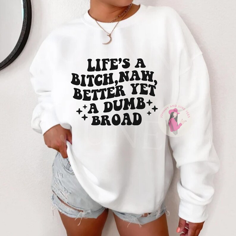 May include: White sweatshirt with the text "LIFE'S A BITCH, NAW, BETTER YET A DUMB BROAD" in black. The sweatshirt has a crew neck and long sleeves. The brand logo is on the right side of the sweatshirt.