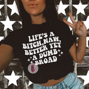 May include: Black crop top with the text "LIFE'S A BITCH, NAH, BETTER YET, A DUMB BROAD" in white, stylized lettering. The shirt features a small circular logo and is worn with blue denim shorts. The background is a black and white star pattern.