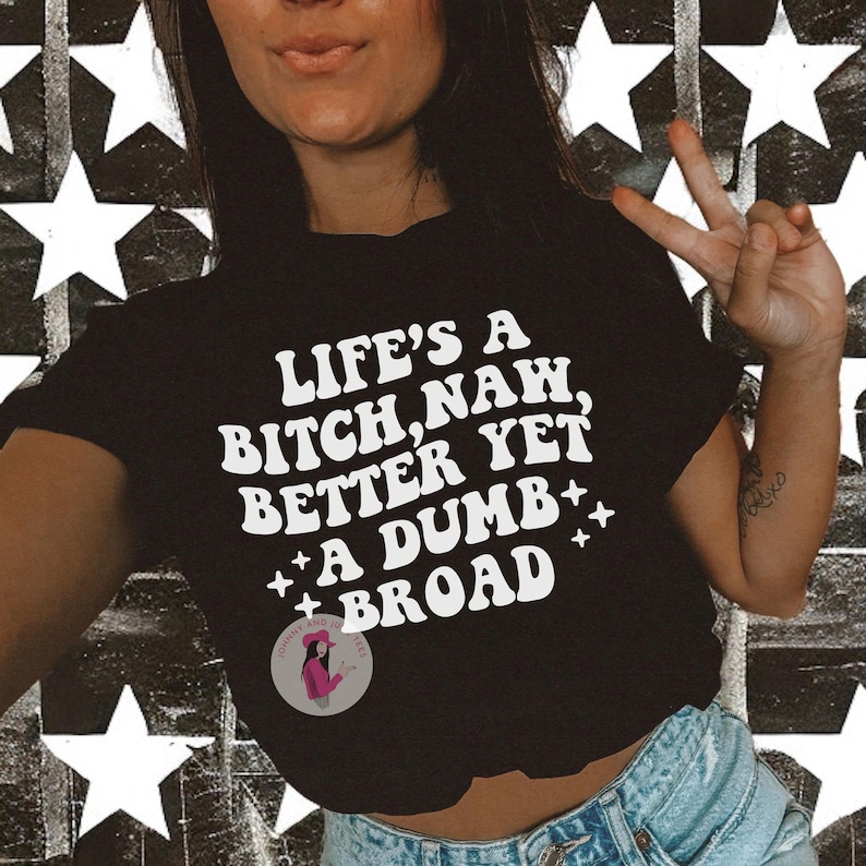 May include: Black crop top with the text "LIFE'S A BITCH, NAH, BETTER YET, A DUMB BROAD" in white, stylized lettering. The shirt features a small circular logo and is worn with blue denim shorts. The background is a black and white star pattern.