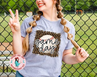 May include: A heather gray t-shirt featuring a baseball graphic with the words "game day" in a script font. The baseball is set against a leopard print background. The shirt has short sleeves.