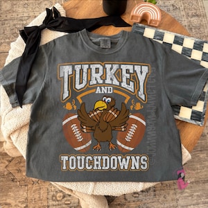May include: Dark gray t-shirt with the words "TURKEY and TOUCHDOWNS" in white and orange lettering. A cartoon turkey with footballs on either side is in the center. The shirt is short-sleeved.