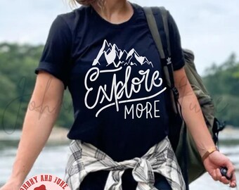 以下が含まれることがあります： 山と「Explore More」という文字が白で描かれた黒いTシャツ。モデルは、腰にチェック柄のシャツを巻き、茶色のベースボールキャップをかぶっている。