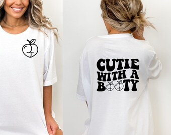 Può includere: Maglietta bianca con una grafica di pesca sul davanti e il testo "Cutie with a Booty" con due grafiche di pesca sul retro.