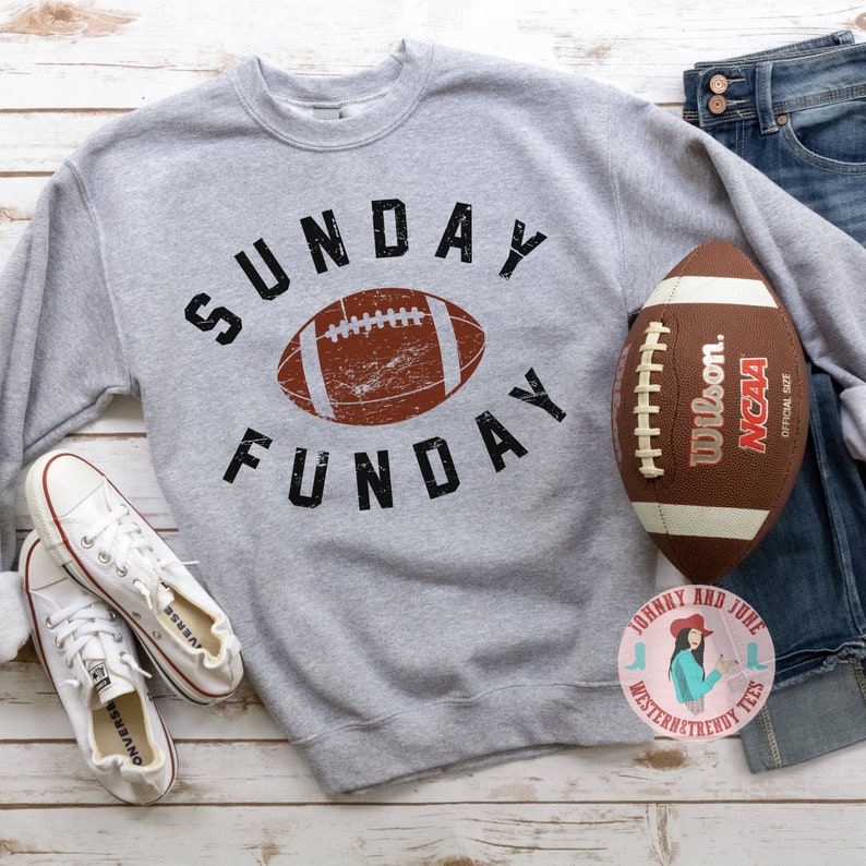 Pu&ograve; includere: Felpa grigia con il testo "SUNDAY FUNDAY" in nero e una grafica di football americano marrone usurata. Un pallone da football americano marrone con lacci bianchi e il testo "Wilson. NCAA OFFICIAL SIZE" si trova nell'angolo in alto a destra dell'immagine.