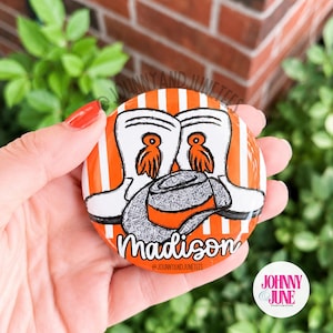 Könnte beinhalten: Ein runder Ansteckbutton mit weiß-orange gestreiftem Hintergrund. Das Motiv zeigt zwei weiße Cowboystiefel mit orangefarbenen Quasten und einen grauen Cowboyhut. Der Name Madison ist in weißer Schreibschrift am unteren Rand aufgedruckt.