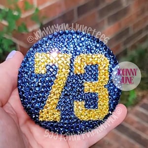 May include: A circular emblem covered in dark blue rhinestones featuring the number 73 in gold rhinestones. The text @johnnyandjunetees is visible in a repeating pattern, and a circular logo with the text JOHNNY JUNE is on the right side.