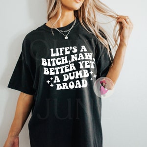May include: Black oversized t-shirt with the text "LIFE'S A BITCH, NAW, BETTER YET + A DUMB + BROAD" in white. The shirt has a crew neck and short sleeves. The shirt is a casual style.