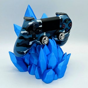 May include: A blue and gray camouflage PlayStation 4 controller is sitting on a blue crystal-shaped stand. The controller has skull-shaped buttons.