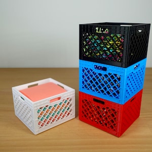May include: Four plastic crates stacked on top of each other. The top crate is black and filled with colorful plastic twist ties. The second crate is blue, the third is red, and the bottom crate is white and contains a pink piece of paper.