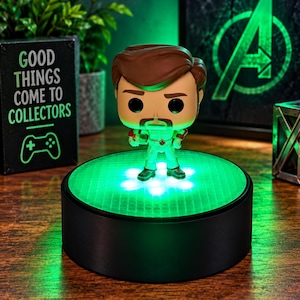 May include: A vinyl collectible figure with brown hair and a mustache, wearing a white jumpsuit with red accents, standing on a circular black display base that emits a bright green light from its textured, translucent top surface.