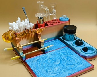 May include: A multi-colored organizer with various compartments and accessories. It includes a glass rig, a lighter, cotton swabs, and tools. The base has a blue, swirled pattern. The organizer is designed to hold smoking accessories.