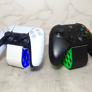 May include: Two video game controllers, one white and one black, are displayed on stands. The white controller has a blue stand and the black controller has a green stand.