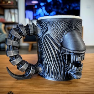 May include: A silver-colored mug with a detailed design of an alien's head and a segmented handle. The mug features a textured surface and a wide open mouth with sharp teeth. The base of the mug has the logo "3DGEEX".