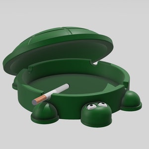 May include: Green turtle-shaped ashtray with a cigarette inside. The ashtray has a lid that is open and the turtle has googly eyes.