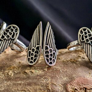 Puede incluir: Tres anillos de plata con diseños de alas de ángel. Las alas están detalladas con un patrón texturizado.