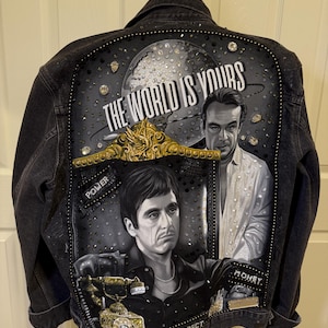 Puede incluir: Una chaqueta vaquera negra con un gran gráfico en la espalda. El gráfico presenta un globo terráqueo con el texto "The World Is Yours" y dos caras de hombres. La chaqueta está decorada con pedrería y tiene las palabras "Power", "Respect" y "Money" escritas en ella.