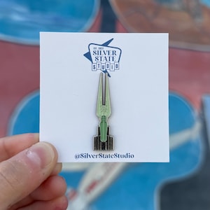 May include: A green and black enamel pin on a white card. The pin is shaped like a stylized weapon. The card has the text "EST. 2023 SILVER STATE STUDIO" and the Instagram handle "@SilverStateStudio".