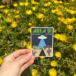 K&ouml;nnte beinhalten: Ein Aufkleber mit einer Cartoon-Illustration einer Stra&szlig;e, die nach Area 51, Nevada f&uuml;hrt. Der Aufkleber zeigt "Area 51" mit "Las Vegas" und "Nevada" darunter. Das Stra&szlig;enschild zeigt "Extraterrestrial Highway".