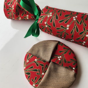 May include: A red and green fabric Christmas cracker with a gold and white mistletoe pattern. The cracker is tied with a green ribbon. A matching fabric round is also shown.