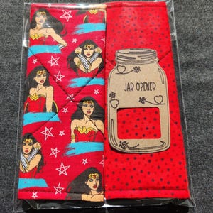 May include: A red jar opener and matching pot holder set. The pot holder features a Wonder Woman design with stars. The jar opener has the words "JAR OPENER" printed on it. The set is packaged in a clear plastic bag.
