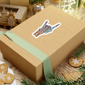Puede incluir: Una caja de regalo marr&oacute;n con una cinta verde y una pegatina de una mano de esqueleto colorida haciendo el signo 'rock on'. La pegatina tiene los nombres de los huesos etiquetados.