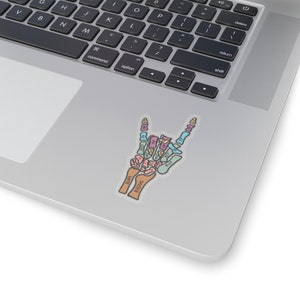 Puede incluir: Una pegatina de una mano de esqueleto haciendo el gesto de rock and roll. Los huesos est&aacute;n etiquetados con sus nombres anat&oacute;micos en colores pastel.