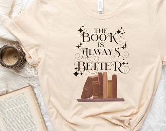 May include: A beige t-shirt with the text "The Book Is Always Better" in black script with a stack of books and gold stars.
