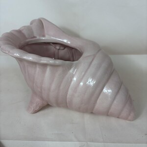 May include: A pale pink ceramic planter shaped like a conch shell. The vessel features a glossy finish, a wide opening at the top, and three small feet at the base to provide stability. The exterior surface has a ribbed texture mimicking a seashell.