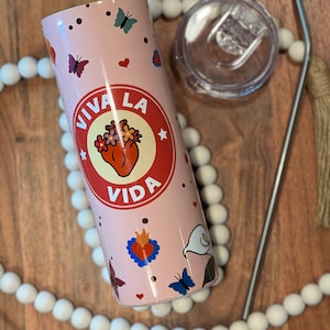 May include: A pink stainless steel tumbler with a white lid and a straw. The tumbler has a design of a heart with flowers and the words "Viva La Vida" in a circle around it. There are also butterflies and hearts on the tumbler.