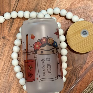 May include: A frosted glass tumbler with a colorful design featuring a woman with a flower crown and the text "viva la vida". The tumbler has a bamboo lid and a clear plastic straw.