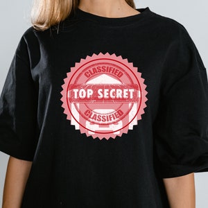 Peut inclure: T-shirt noir avec un tampon rouge et blanc "CLASSIFIED TOP SECRET".