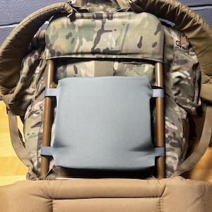 May include: A camouflage backpack with a grey back support and brown wooden frame. The backpack has padded shoulder straps and a top handle. The fabric has a mix of green, brown, and tan colours.