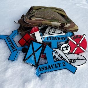 Op de afbeelding: Een verzameling blauwe en rode militaire markers met verschillende tekst, waaronder "Support", "Assault", "CP" en "OBJ". De markers zijn verspreid over een wit oppervlak, waarschijnlijk sneeuw.