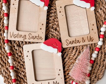 May include: Three wooden photo frames with Santa hats, the text "Visiting Santa", "Santa & Us", and "Santa & Me". The frames are on a woven mat with a red and white beaded garland and a pink Christmas tree.
