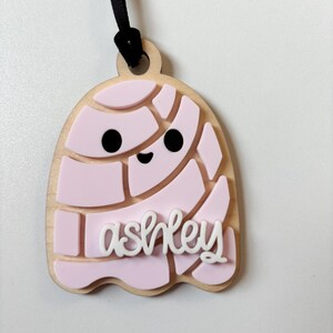 May include: A wooden tag with a light pink igloo design and the name "ashley" in white script. The tag has a black ribbon for hanging.