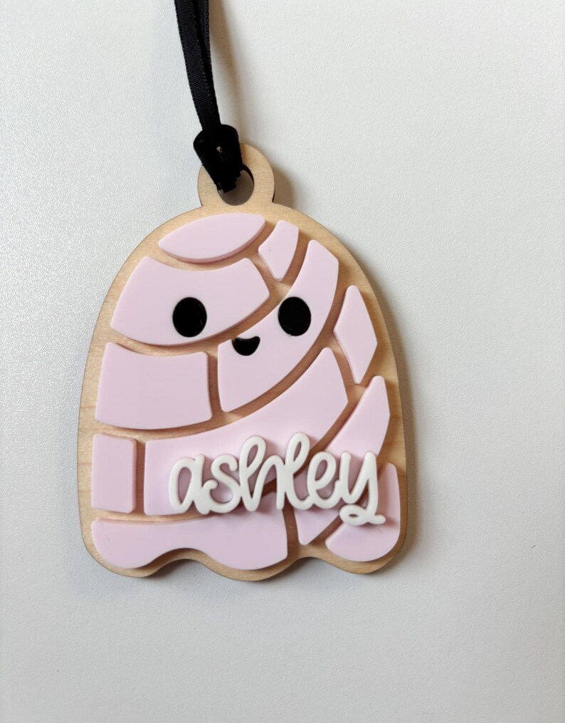 May include: A wooden tag with a light pink igloo design and the name "ashley" in white script. The tag has a black ribbon for hanging.