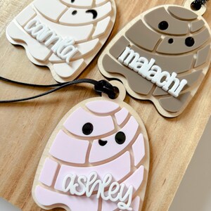 May include: Three personalized laser-cut ornaments in the shape of a concha pastry. One is light beige with the name "camila", another is brown with "malachi", and the last is pink with "ashley". Each has a black string for hanging.