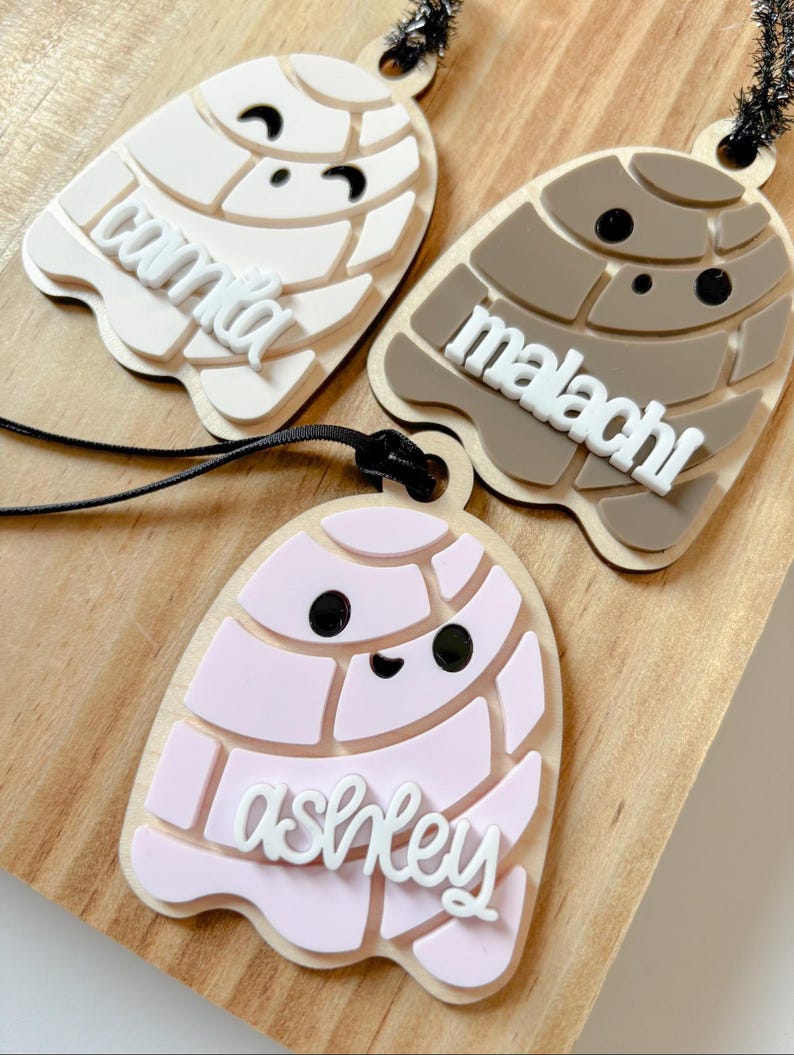 May include: Three personalized laser-cut ornaments in the shape of a concha pastry. One is light beige with the name "camila", another is brown with "malachi", and the last is pink with "ashley". Each has a black string for hanging.