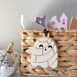 May include: A white ghost-shaped ornament with a black cord hangs in front of a woven basket. The ghost has a smiling face and a segmented design. A silver disco ball pumpkin and decorative houses are also in the image.