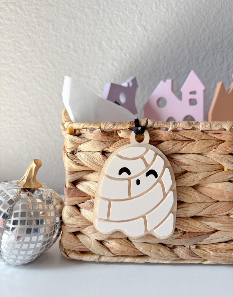May include: A white ghost-shaped ornament with a black cord hangs in front of a woven basket. The ghost has a smiling face and a segmented design. A silver disco ball pumpkin and decorative houses are also in the image.