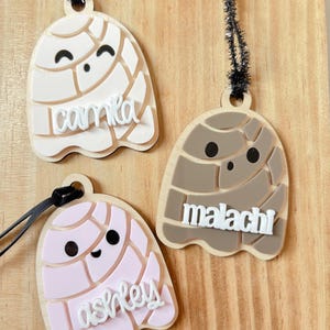 May include: Three wooden concha-shaped ornaments in cream, pink, and brown, each with a name in white script. The ornaments have black eyes and a black string for hanging. The names are Camila, Ashley, and Malachi.