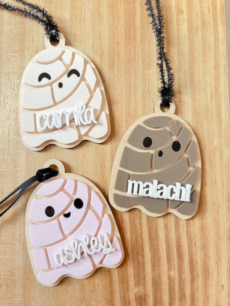 May include: Three wooden concha-shaped ornaments in cream, pink, and brown, each with a name in white script. The ornaments have black eyes and a black string for hanging. The names are Camila, Ashley, and Malachi.