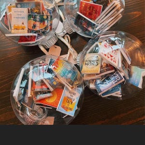 May include: Three clear glass ornaments filled with miniature book covers. The ornaments are hanging from a white string. The book covers are a variety of colors and feature titles such as "Between Two Kingdoms", "Hello Beautiful", "Maybe You Should Talk to Someone", "The Couple Next Door", and "Fourth Wing".