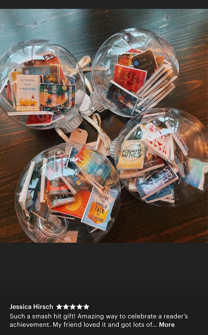 May include: Three clear glass ornaments filled with miniature book covers. The ornaments are hanging from a white string. The book covers are a variety of colors and feature titles such as "Between Two Kingdoms", "Hello Beautiful", "Maybe You Should Talk to Someone", "The Couple Next Door", and "Fourth Wing".