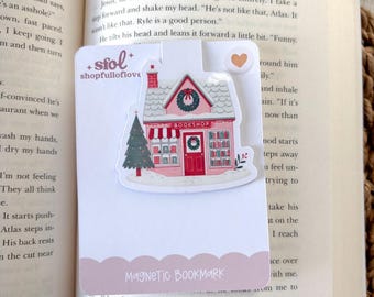 May include: A pink and white magnetic bookmark shaped like a bookstore, with a Christmas tree and wreath. The bookmark is placed in a book with the text "Magnetic Bookmark" and "BOOKSHOP". The shopfulloflove logo is also visible.