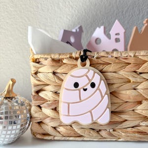 May include: A light-colored, ghost-shaped pendant with a pink design and black eyes and mouth hangs from a black cord. The pendant is attached to a woven basket. A silver disco ball pumpkin is to the left.